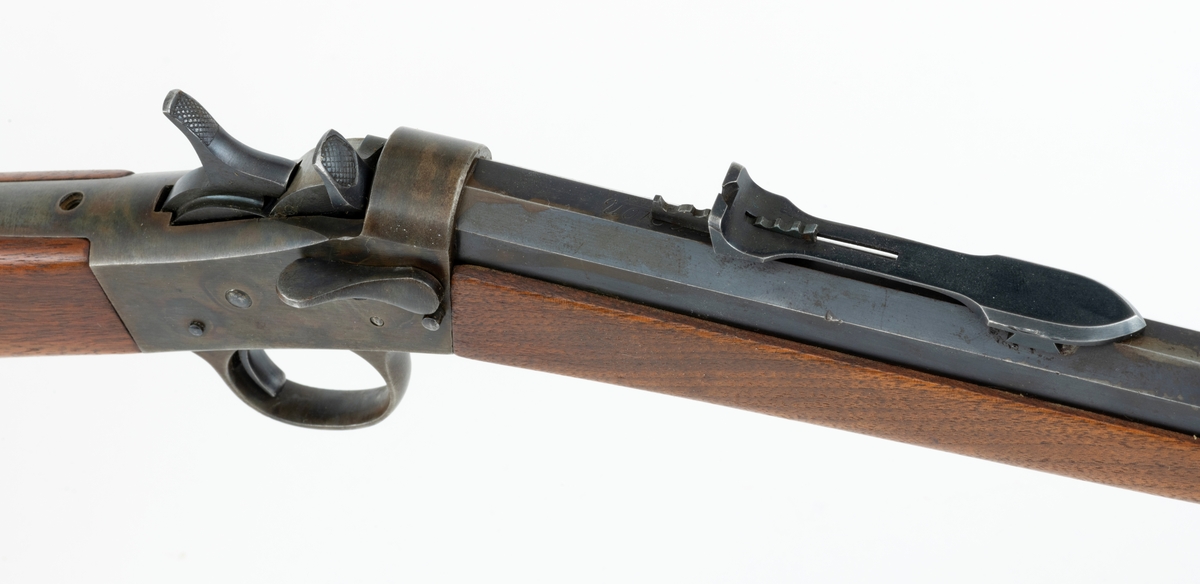 Tillegg innskrift: Remington TRADE MARK MODEL 4 25-10 R. F. Geværet har brunt, oljebehandlet treverk. Framstillingsnummeret er J 304870. Laget ca. 1870. Av størrelse kan geværet se ut som ei salongrifle, men kaliberet, 25-10, er litt større enn på Krag. R. F. står for Rand Fire (randtenning. ) 10-tallet står for 10 gram svartkrutt. Dvs. ca. 4 gram. Dette er en meget kraftig ladning, nesten like mye som på den store Remington. Det må være en av de raskeste svartkruttpatroner som er laget.