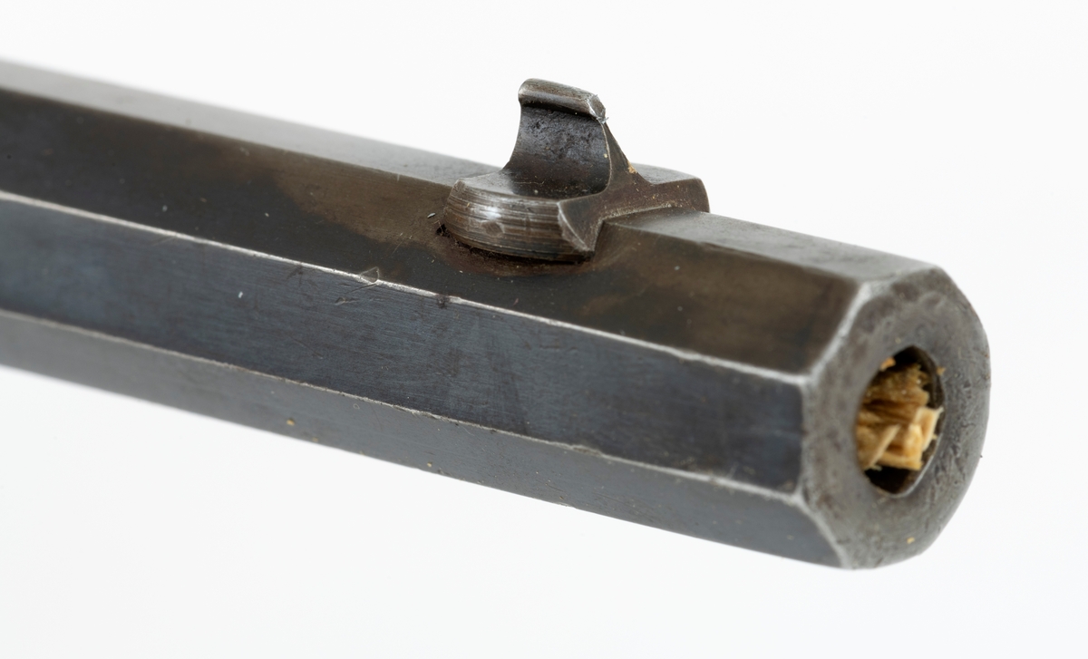 Tillegg innskrift: Remington TRADE MARK MODEL 4 25-10 R. F. Geværet har brunt, oljebehandlet treverk. Framstillingsnummeret er J 304870. Laget ca. 1870. Av størrelse kan geværet se ut som ei salongrifle, men kaliberet, 25-10, er litt større enn på Krag. R. F. står for Rand Fire (randtenning. ) 10-tallet står for 10 gram svartkrutt. Dvs. ca. 4 gram. Dette er en meget kraftig ladning, nesten like mye som på den store Remington. Det må være en av de raskeste svartkruttpatroner som er laget.