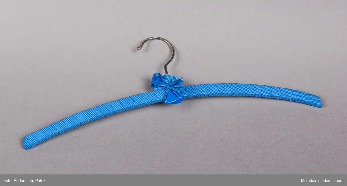 Klädgalge av den enkla typen att hänga skjorta eller blus på. Klädd med omlindat plastband som är pepitamönstrat i blå grundton. Knuten rosett vid hängkroken, som är av stål.