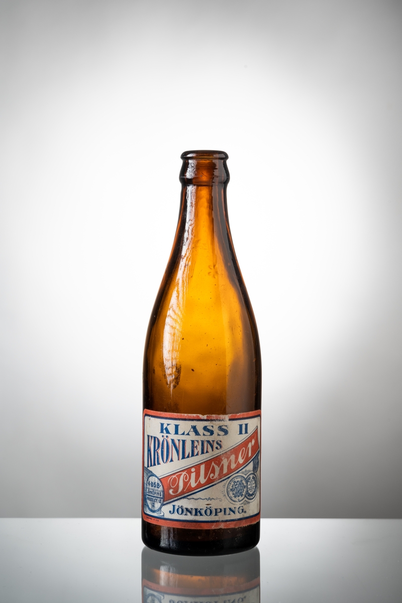 Ölflaskor, 3 st tomma, av brunt glas med påklistrad pappersetikett. Vit etikett med rött och blått tryck, text: "KLASS II KRÖNLEINS Pilsner JÖNKÖPING". På flaskans hals präglat "H" i glaset och tre blomformer. Under botten är präglat siffran 2, 7 respektive 8.

Se vidare Historik.