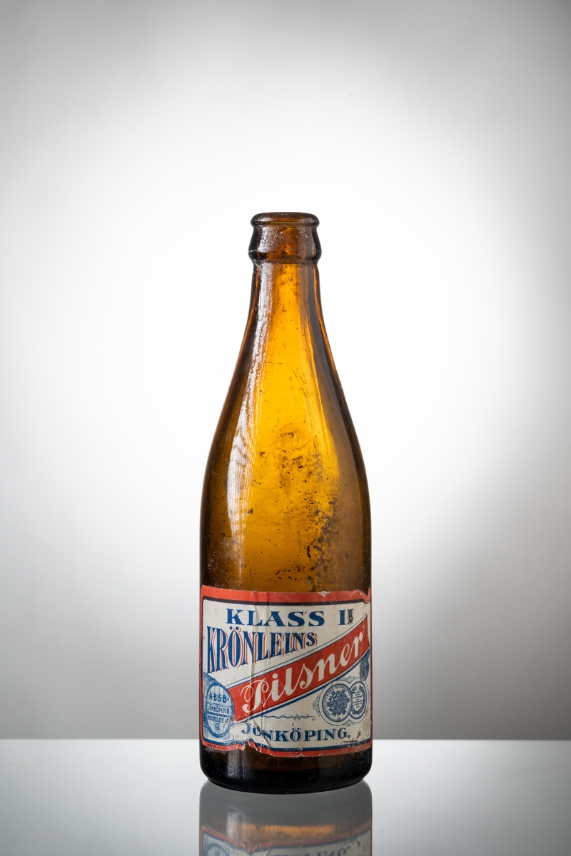 Ölflaskor, 3 st tomma, av brunt glas med påklistrad pappersetikett. Vit etikett med rött och blått tryck, text: "KLASS II KRÖNLEINS Pilsner JÖNKÖPING". På flaskans hals präglat "H" i glaset och tre blomformer. Under botten är präglat siffran 2, 7 respektive 8.

Se vidare Historik.