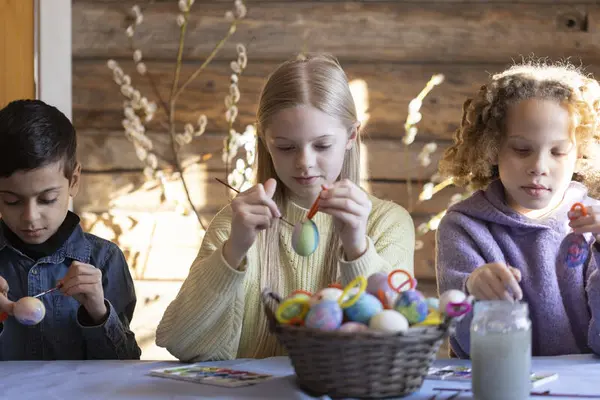 Tre barn sitter ved et bord og maler påskeegg på Norsk Folkemuseum. De bruker pensler og maling for å dekorere eggene, som ligger i en kurv foran dem.