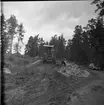 Vägarbete mellan Bunnström och Östanå 1960. Två män står i en stor stenhög. Marken är lerigt. En skogsmaskin står bakom dem.