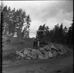 Vägarbete mellan Bunnström och Östanå 1960. Två män står i en stor stenhög. Marken är lerigt. En skogsmaskin står bakom dem.