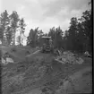 Vägarbete mellan Bunnström och Östanå 1960. Två män står i en stor stenhög. Marken är lerigt. En skogsmaskin står bakom dem.