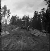 Vägarbete mellan Bunnström och Östanå 1960. Två män står i en stor stenhög. Marken är lerigt. En skogsmaskin står bakom dem.