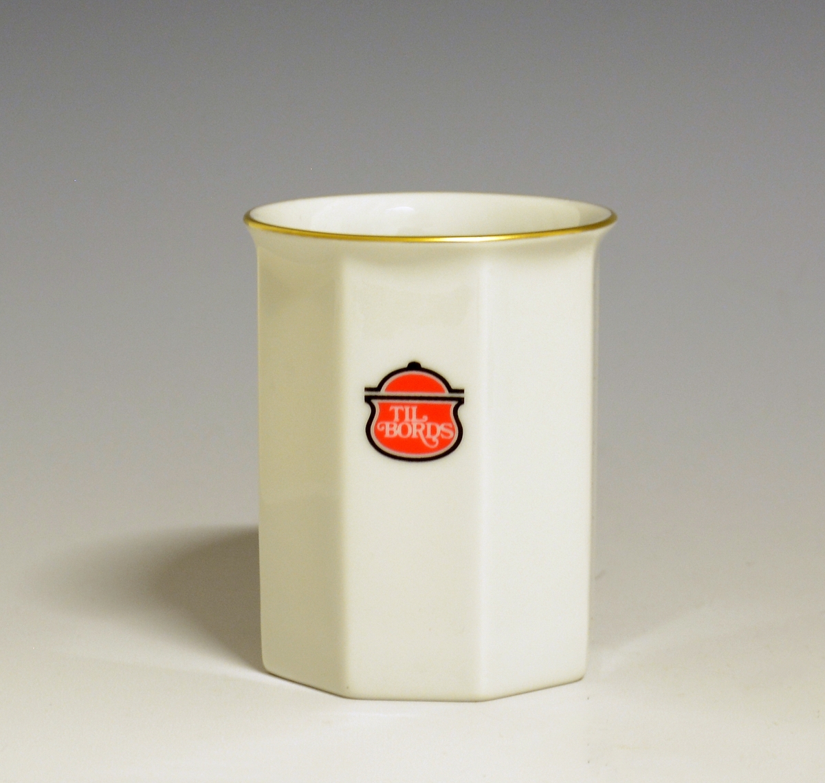 Mangekantet kopp i porselen med kantstrek i gull og logo i form av en rød gryte med lokk og påskriften "Til Bords". Hvit glasur.
Modell: Octavia, tegnet av Grete Rønning i 1977.