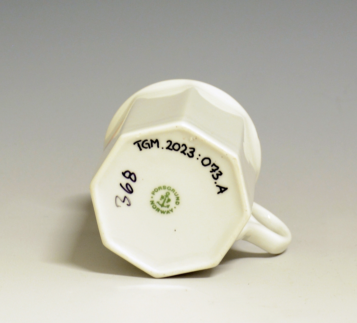 Mangekantet kopp i porselen med kantstrek i gull og logo i form av en rød gryte med lokk og påskriften "Til Bords". Hvit glasur.
Modell: Octavia, tegnet av Grete Rønning i 1977.