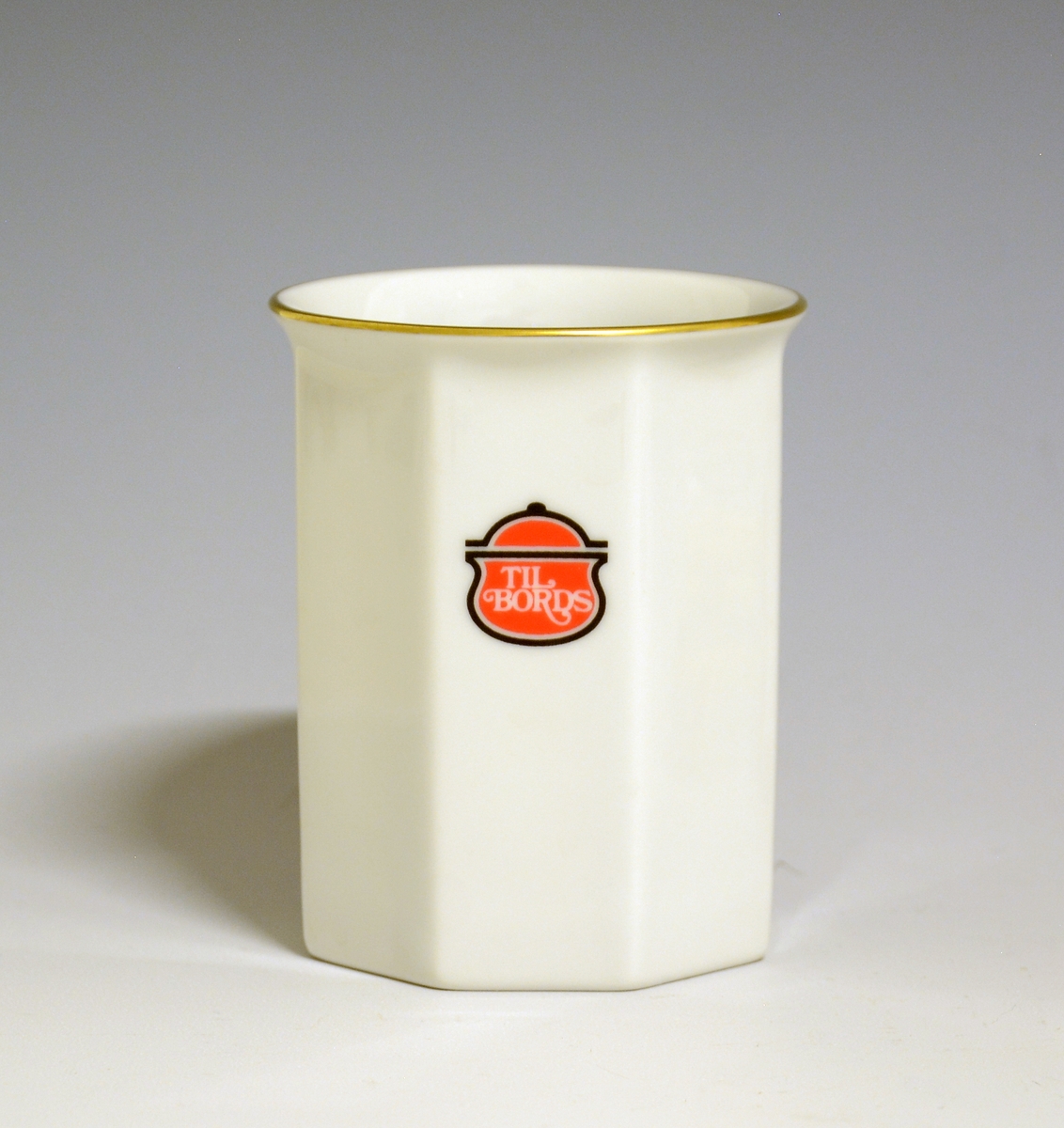 Mangekantet kopp i porselen med kantstrek i gull og logo i form av en rød gryte med lokk og påskriften "Til Bords". Hvit glasur.
Modell: Octavia, tegnet av Grete Rønning i 1977.
