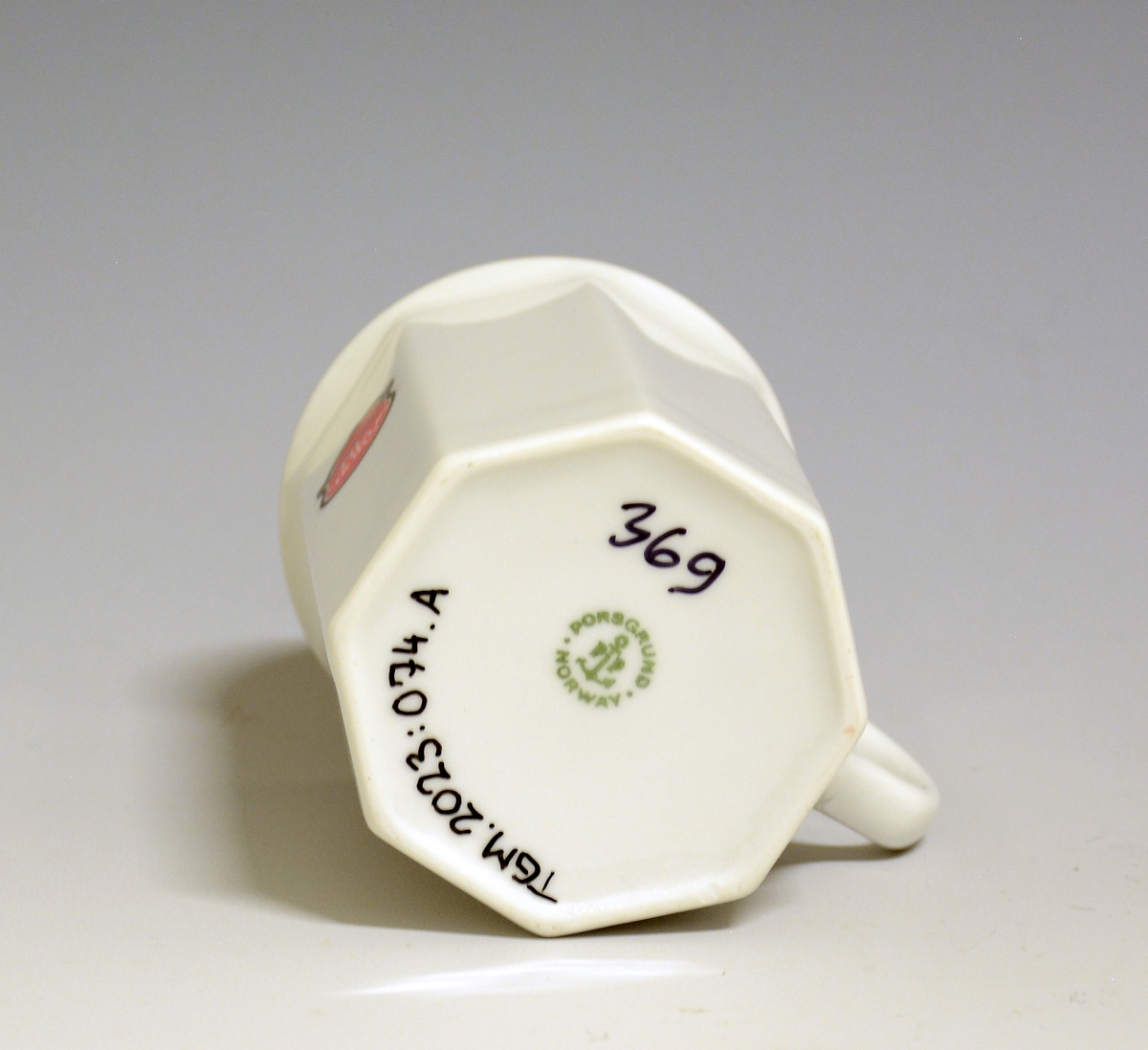 Mangekantet kopp i porselen med kantstrek i gull og logo i form av en rød gryte med lokk og påskriften "Til Bords". Hvit glasur.
Modell: Octavia, tegnet av Grete Rønning i 1977.
