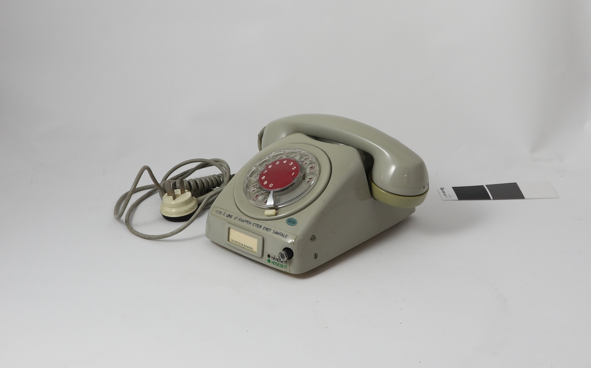 Grått telefonapparat av bakelitt med tilnærmet kvadratisk bunn og buet deksel med sirkulær tallskive.
