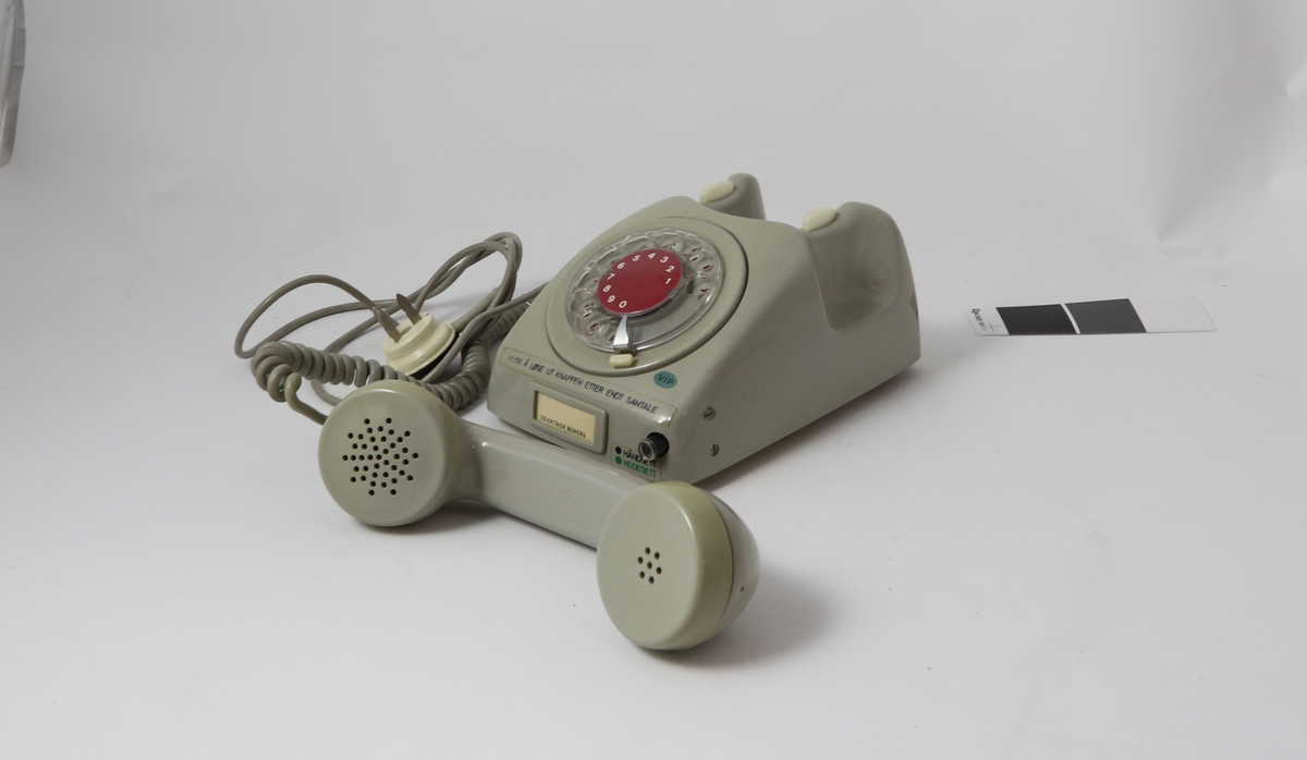 Grått telefonapparat av bakelitt med tilnærmet kvadratisk bunn og buet deksel med sirkulær tallskive.
