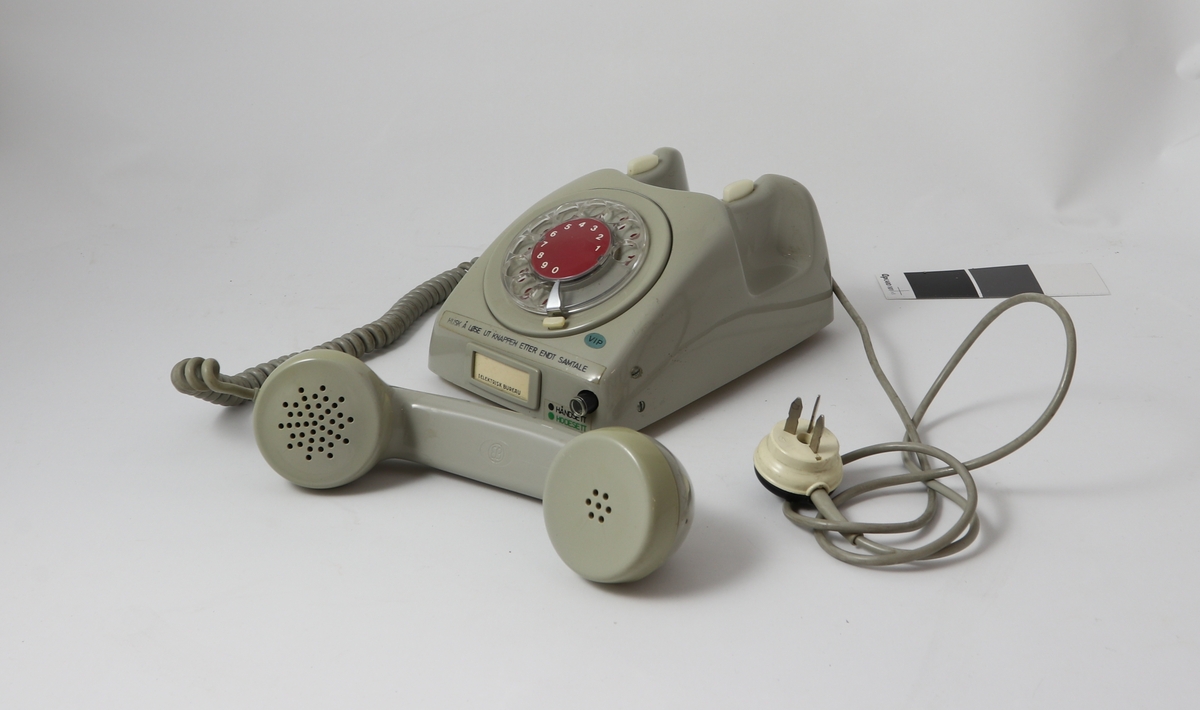 Grått telefonapparat av bakelitt med tilnærmet kvadratisk bunn og buet deksel med sirkulær tallskive.
