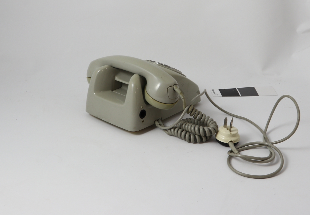 Grått telefonapparat av bakelitt med tilnærmet kvadratisk bunn og buet deksel med sirkulær tallskive.
