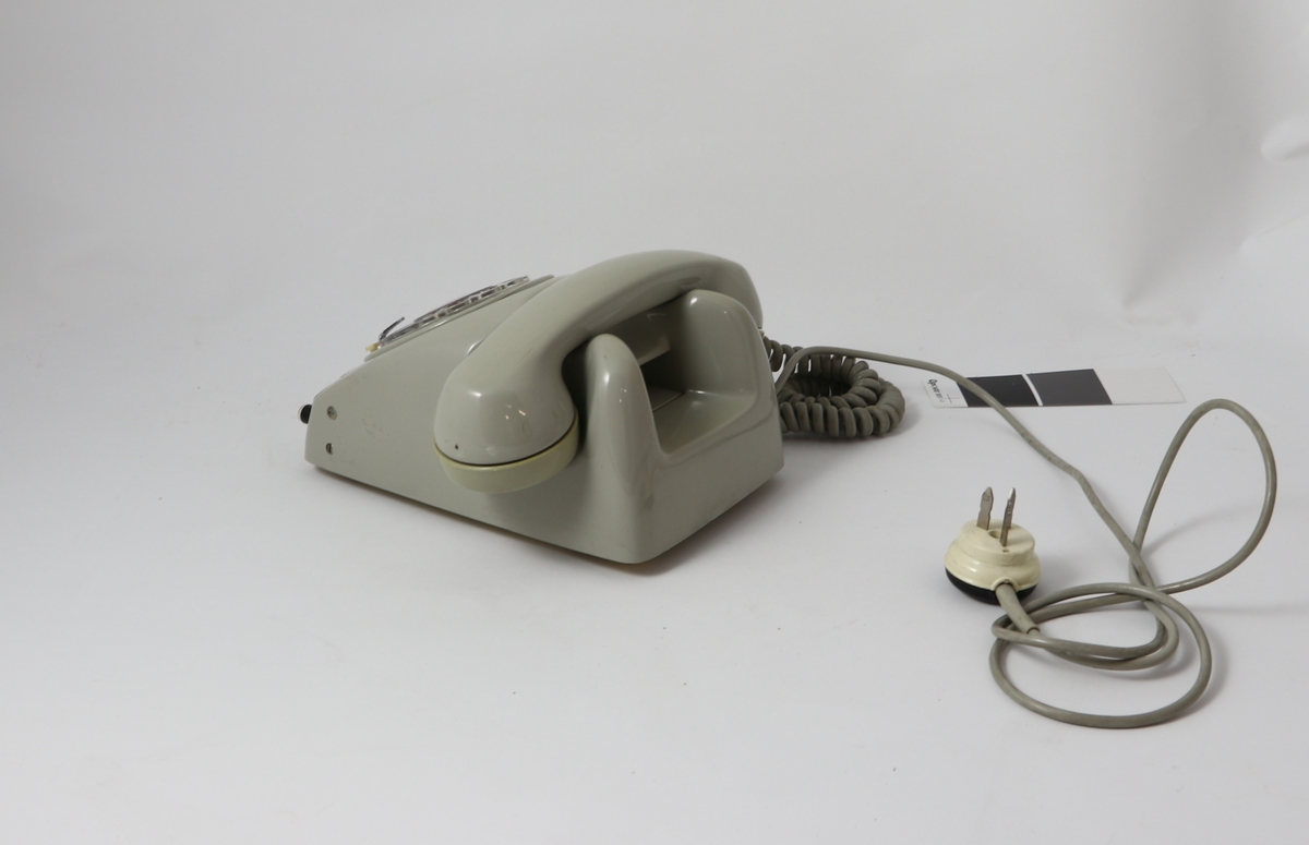 Grått telefonapparat av bakelitt med tilnærmet kvadratisk bunn og buet deksel med sirkulær tallskive.

