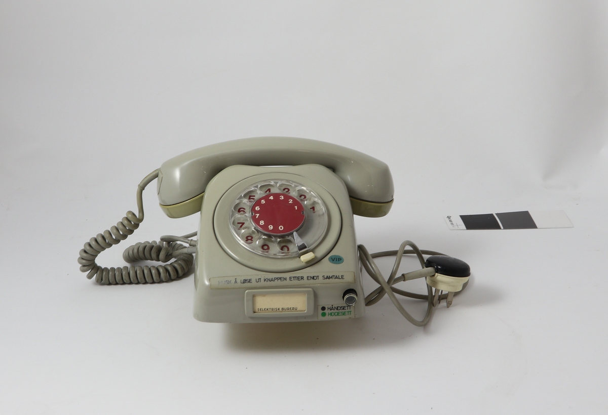 Grått telefonapparat av bakelitt med tilnærmet kvadratisk bunn og buet deksel med sirkulær tallskive.
