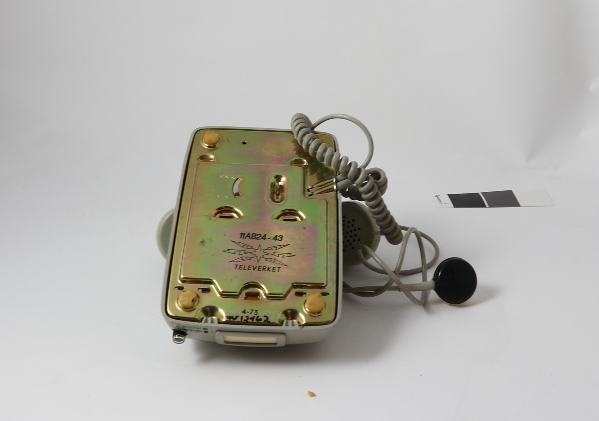 Grått telefonapparat av bakelitt med tilnærmet kvadratisk bunn og buet deksel med sirkulær tallskive.
