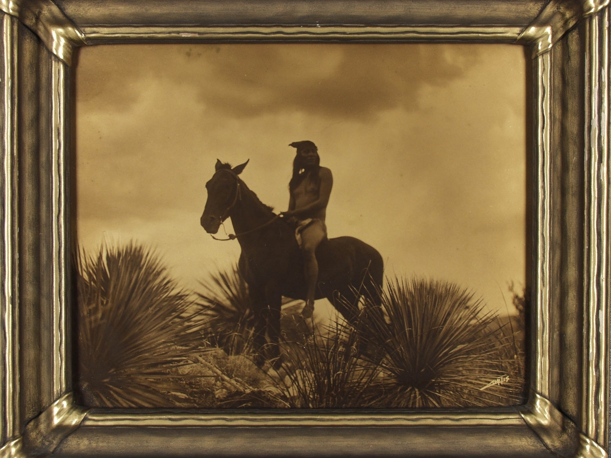 Apache til hest, med vegetasjon i forgrunnen.