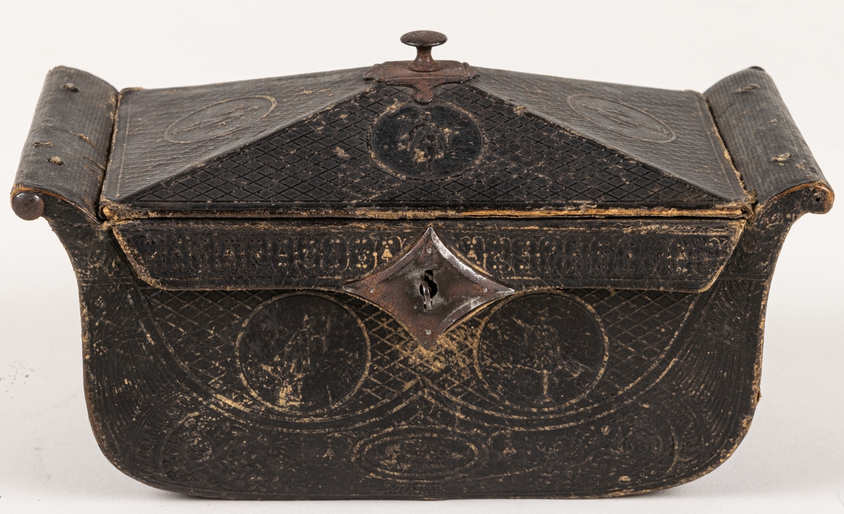 Ask, av papp, svart, med pressat empiremönster: rutor, blomster, medaljonger /runda med gudafigurer/ mm. Ytterformen är sarkofagartad, med uppåt-utåtböjda gavelstycken och lock av pyramidtakform, med metallknopp på mitten. På lockets insida är fästad en penningbörsliknande ficka.