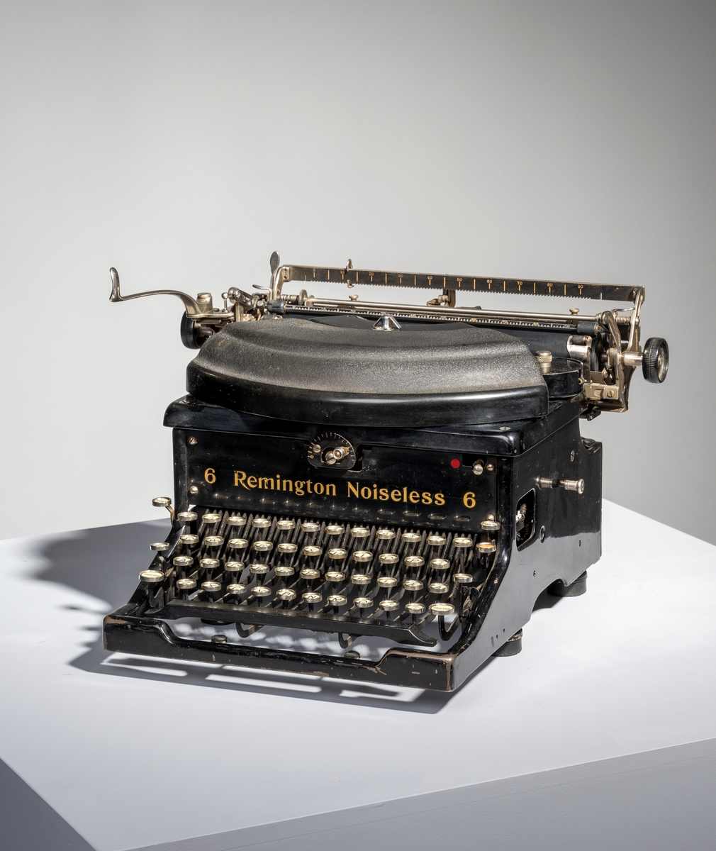 En skrivemaskin av typen "Remington Noiseless 6". Den er av svartlakkert metall med påført tekst i gullfarge. Den står på fire runde bein av svart gummi. Typearmene er festet i vifteform og over dem er det et deksle av svart bakelitt. På hver side av vifteformen er det runde holdere med deksel for å sette i fargebånd. I bakkant på maskinen er det stativ for å sett i papir. Papiret justeres med svarte plasthjul montert på hver ende av stativet. På venstre side er det et håndtak for å kunne skyve papiret opp to hakk. På stativet er det også tre linjaler/målestokker, to i bakkant og en i front. 
I front på skrivemaskinen er det tastatur. Det er tre rader med bokstaver med "dobbel shift" for henholdsvis store og små bokstaver. Nederst til høyre (nederste bokstavrekke) er det to taster med henholdsvis "+." og "-,". Øverst på tastaturet er det en rad med tall fra 1-0 samt ulike tegn som 
$ - % - § og så videre. Ytterst til venstre er det tre taster for henholdsvis "Tilbage", "Holder" og "Skifter". Ytterst til høyre er det fire taster for henholdsvis "Marginal udløsn.", "TAB", "Holder" og "SKifter. Nederst er det en avlang mellomromstast. Alle tastene er hvite med svart skrift unntatt "TAB" som er rød med hvit skrift og mellomromstasten som er svart. Til skrivemaskinen hørere det til et firkantet underlag av grønn filt og et trekk sydd av brunt kunststoff/skai.