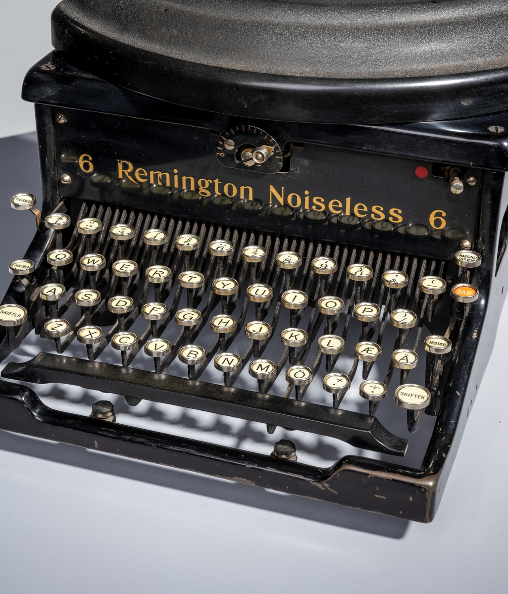 En skrivemaskin av typen "Remington Noiseless 6". Den er av svartlakkert metall med påført tekst i gullfarge. Den står på fire runde bein av svart gummi. Typearmene er festet i vifteform og over dem er det et deksle av svart bakelitt. På hver side av vifteformen er det runde holdere med deksel for å sette i fargebånd. I bakkant på maskinen er det stativ for å sett i papir. Papiret justeres med svarte plasthjul montert på hver ende av stativet. På venstre side er det et håndtak for å kunne skyve papiret opp to hakk. På stativet er det også tre linjaler/målestokker, to i bakkant og en i front. 
I front på skrivemaskinen er det tastatur. Det er tre rader med bokstaver med "dobbel shift" for henholdsvis store og små bokstaver. Nederst til høyre (nederste bokstavrekke) er det to taster med henholdsvis "+." og "-,". Øverst på tastaturet er det en rad med tall fra 1-0 samt ulike tegn som 
$ - % - § og så videre. Ytterst til venstre er det tre taster for henholdsvis "Tilbage", "Holder" og "Skifter". Ytterst til høyre er det fire taster for henholdsvis "Marginal udløsn.", "TAB", "Holder" og "SKifter. Nederst er det en avlang mellomromstast. Alle tastene er hvite med svart skrift unntatt "TAB" som er rød med hvit skrift og mellomromstasten som er svart. Til skrivemaskinen hørere det til et firkantet underlag av grønn filt og et trekk sydd av brunt kunststoff/skai.