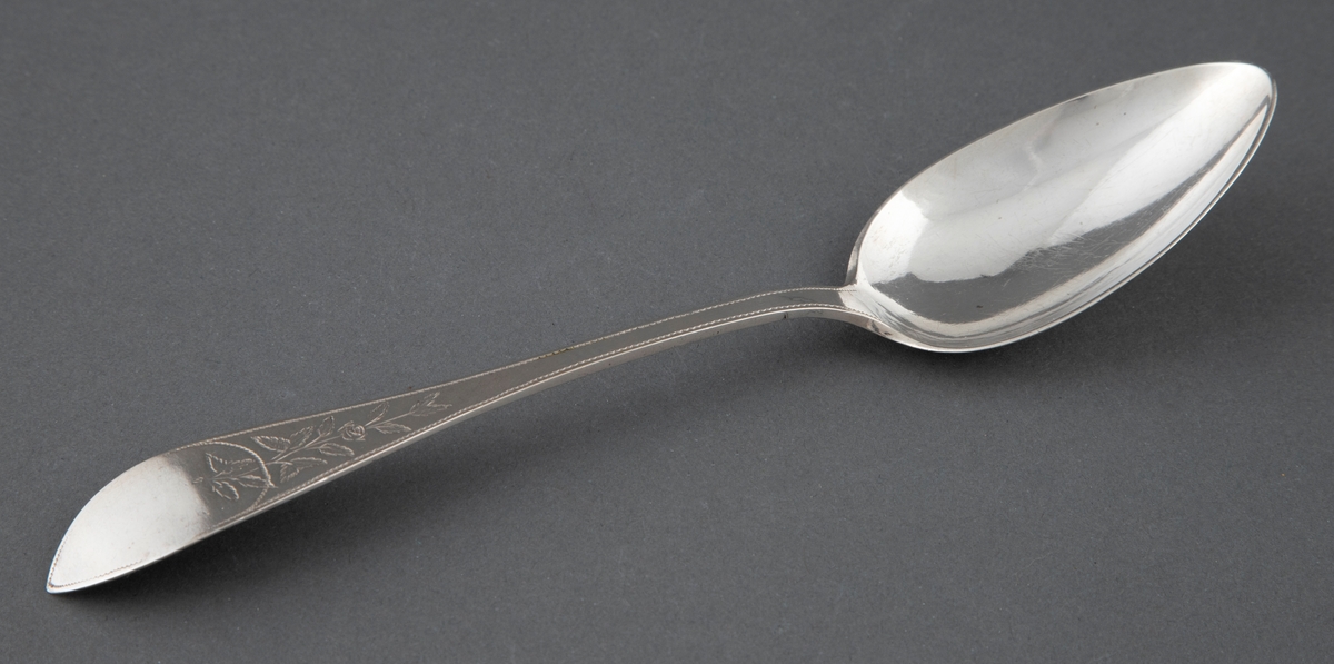 En av seks like spiseskjeer i sølv. Skjebladet er spisst og eggformet. Flat skaft med spiss avslutning. Enkel gravyr med blomsterranke og prikkebord.