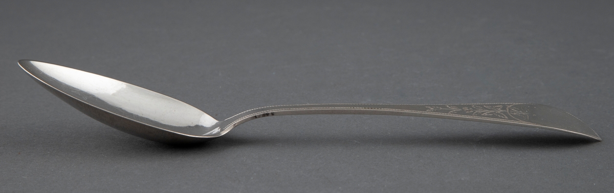 En av seks like spiseskjeer i sølv. Skjebladet er spisst og eggformet. Flat skaft med spiss avslutning. Enkel gravyr med blomsterranke og prikkebord.