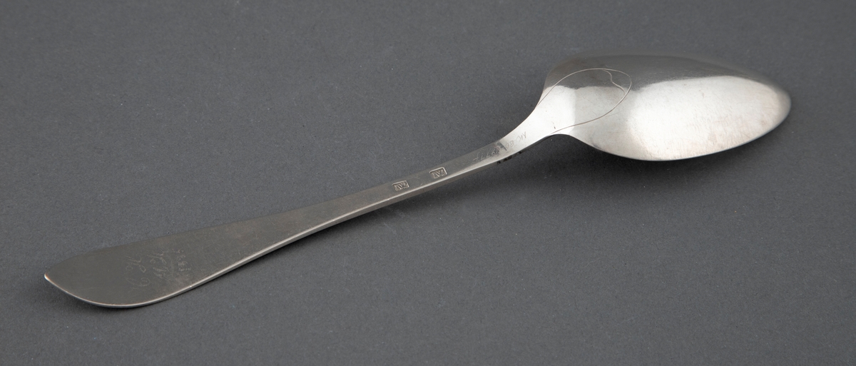 En av seks like spiseskjeer i sølv. Skjebladet er spisst og eggformet. Flat skaft med spiss avslutning. Enkel gravyr med blomsterranke og prikkebord.