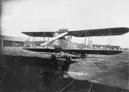 Flygplan FVM S 21 uppställt på ett fält, omkring 1922-1926. I bakgrunden syns en tranformator och ett stall.