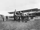 Ambulansflygplan Bréguet XIV Tbis, civilregistrerat S-ASAA, står framför hangarer på ett flygfält omkring 1924. Planet köptes in till Röda Korset från Frankrike, var i tjänst 1924 -1926 med Boden som bas. Folksamling kring flygplanet.