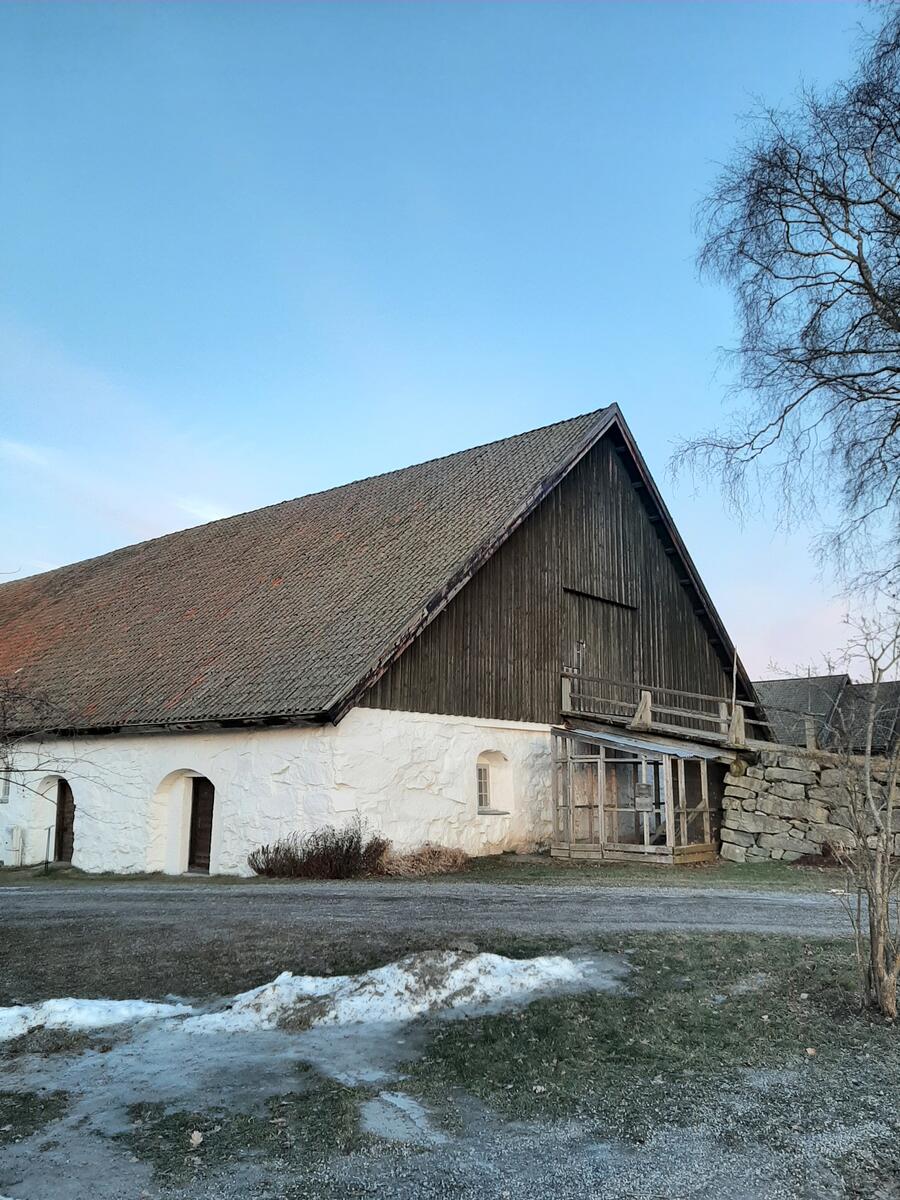 Bildet viser fremsiden av det hvitt steinfjøs. Fjøset er delt i to, der det er hvitt på undersiden og brun plank på oversiden av det hvite.