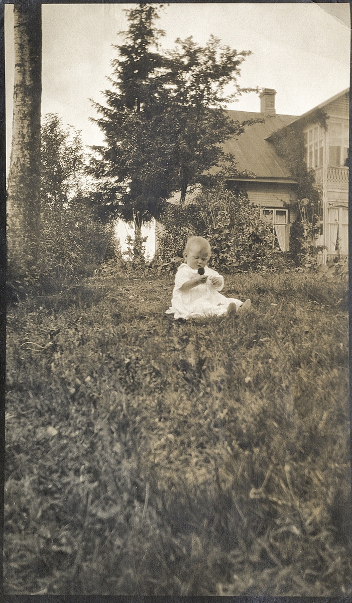 En baby sitter på gräsmattan i en trädgård. I bakgrunden skymtar ett bostadshus med glasveranda.