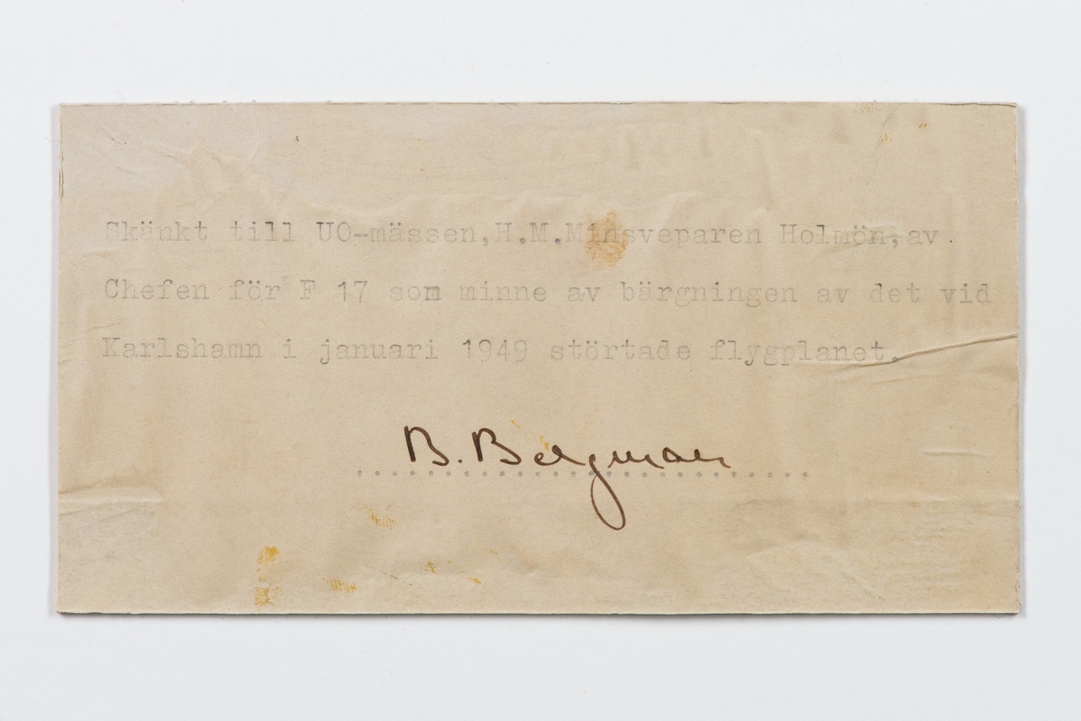 Fotografi inom ram av mahogny. På ramen en bricka av nysilver med inskriptionen: F 17 Bärgning av flp. T 18 B jan. 1949.
På tavlans baksida följande text: Skänkt till gunrummet H.M Minsveparen Holmön av chefen för F17 som minne av bärgningen av det vid Karlshamn i januari 1949 störtade flygplanet. B. Bergman F L. Texten på baksidan som här utgöres av: Skänkt till UO-mässen.
Neg.nr 4928.