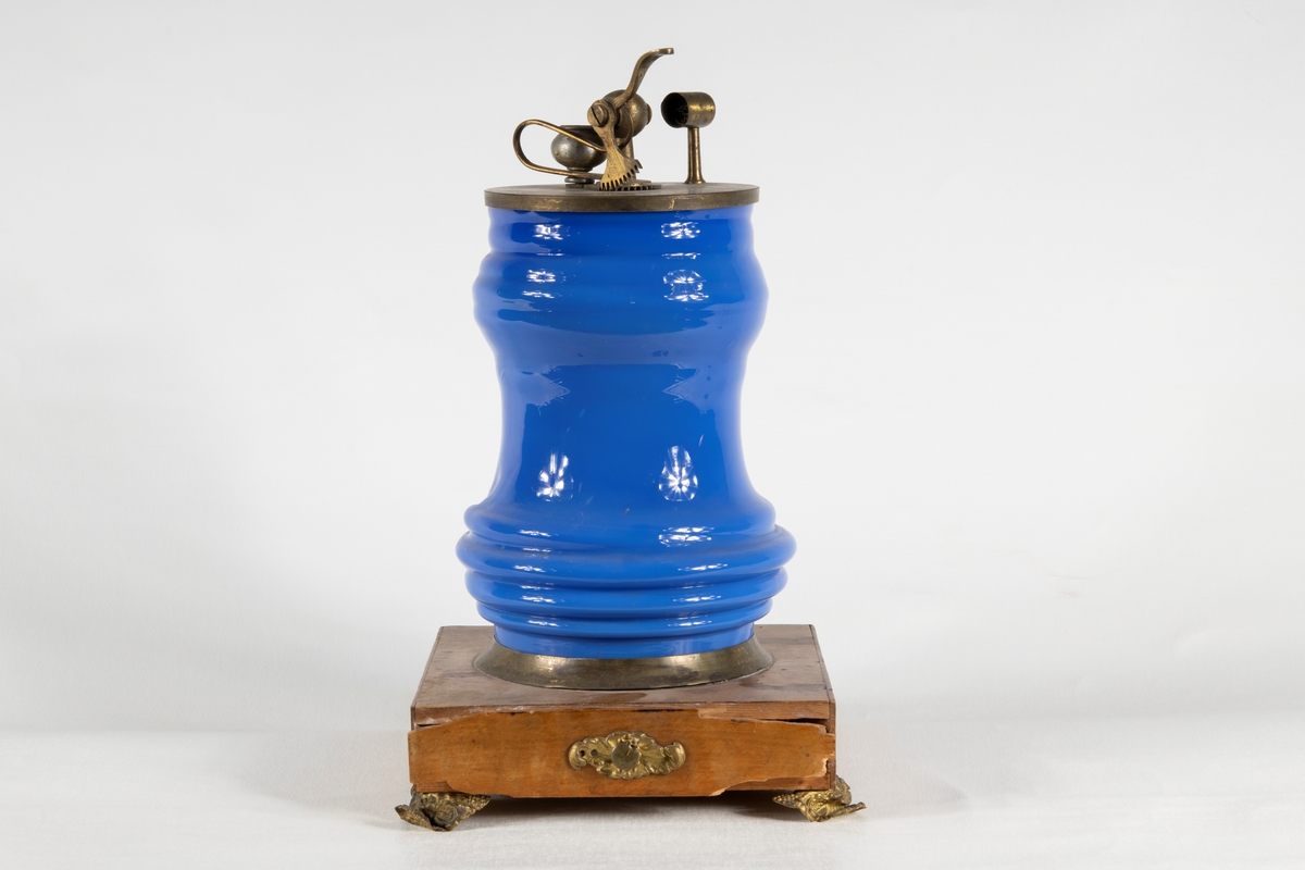 Ett Döbereinerskt elddon på träsockel, med fyra metallfötter. Elddonet består av fyra delar; En glaskupa i blått handblåst glas, träsockel med metallsarg och metallfötter samt utdragsfack. Glaskupa i genomskinligt glas samt mekanik för antändning i metall, samt en platinasvamp med upphängningstång i zink och kolv.