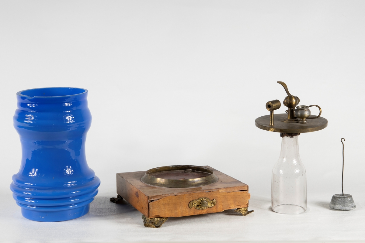 Ett Döbereinerskt elddon på träsockel, med fyra metallfötter. Elddonet består av fyra delar; En glaskupa i blått handblåst glas, träsockel med metallsarg och metallfötter samt utdragsfack. Glaskupa i genomskinligt glas samt mekanik för antändning i metall, samt en platinasvamp med upphängningstång i zink och kolv.