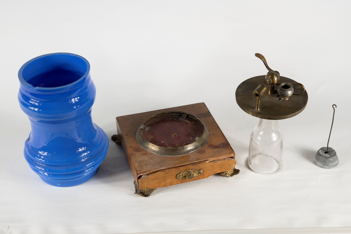 Ett Döbereinerskt elddon på träsockel, med fyra metallfötter. Elddonet består av fyra delar; En glaskupa i blått handblåst glas, träsockel med metallsarg och metallfötter samt utdragsfack. Glaskupa i genomskinligt glas samt mekanik för antändning i metall, samt en platinasvamp med upphängningstång i zink och kolv.