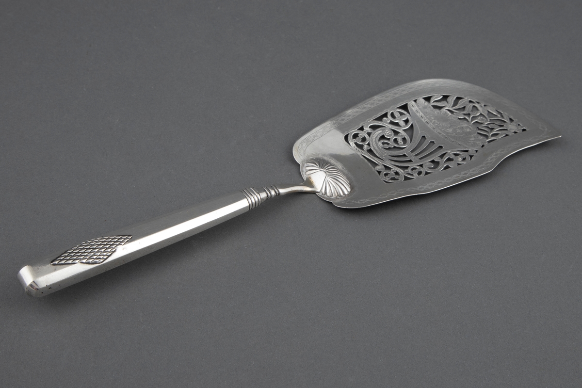 Fiskespade i sølv med gjennombrutt blad og blomsterhorn-motiv. Muslingaktig dekor ved bladroten og mangekantet skaft med rombedekor ved skaftenden.