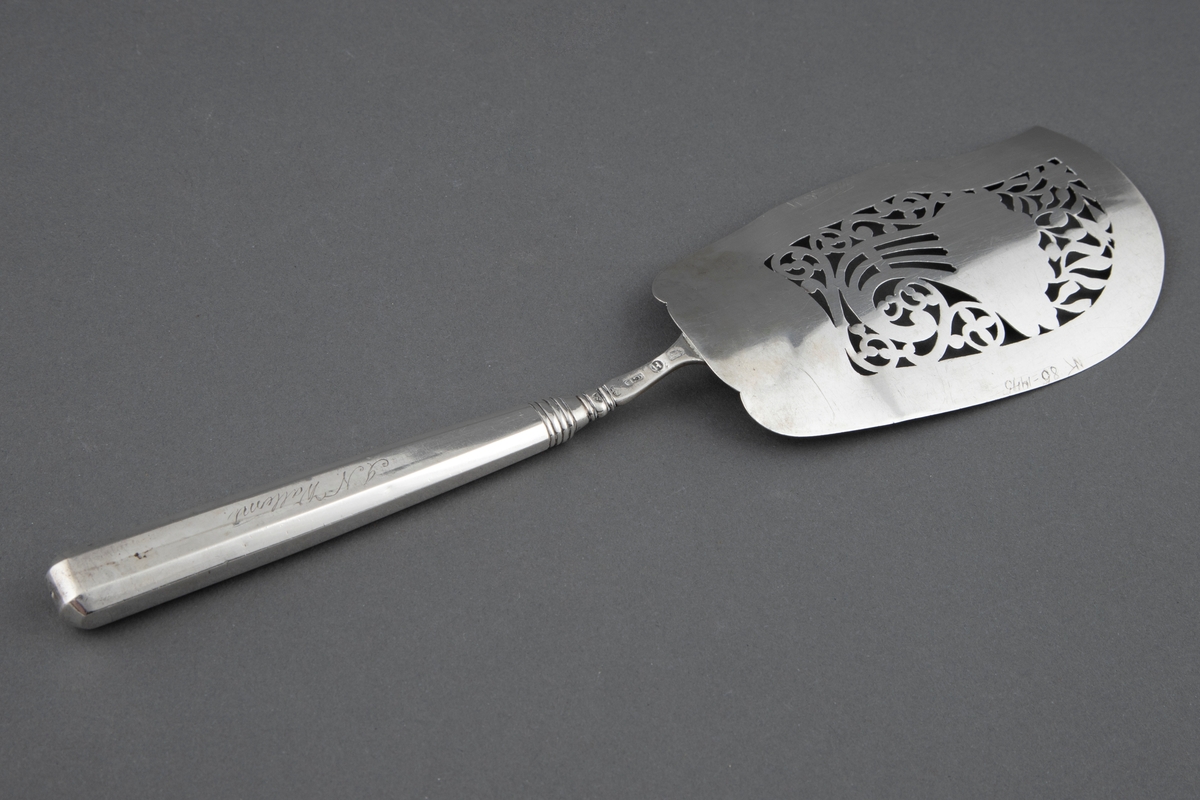 Fiskespade i sølv med gjennombrutt blad og blomsterhorn-motiv. Muslingaktig dekor ved bladroten og mangekantet skaft med rombedekor ved skaftenden.