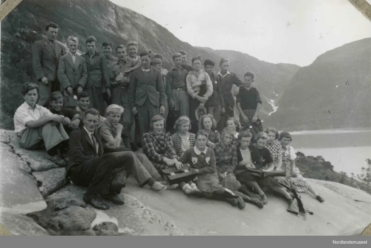 En stor gruppe unge mennesker som sitter på et glatt berg. I bakgrunnen ser vi et vann/sjø. fjell og et fossefall. Tekst i album: Kl. 2 1938 med klasseforstander.