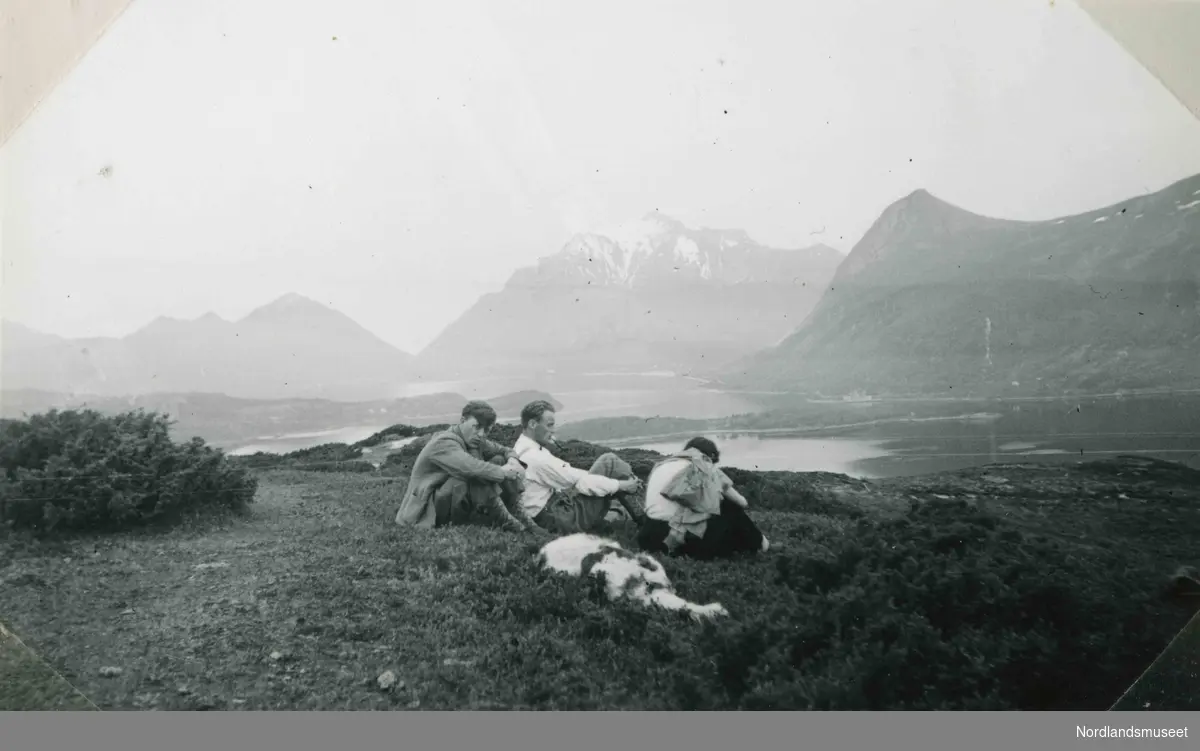 Tre menn sitter på en høyde. Den ene har pipe i munnen. I bakgrunnen ser vi fjell og fjord. Tekst i album: På tur Glomfjorden, utsikt mot Gildeskål.