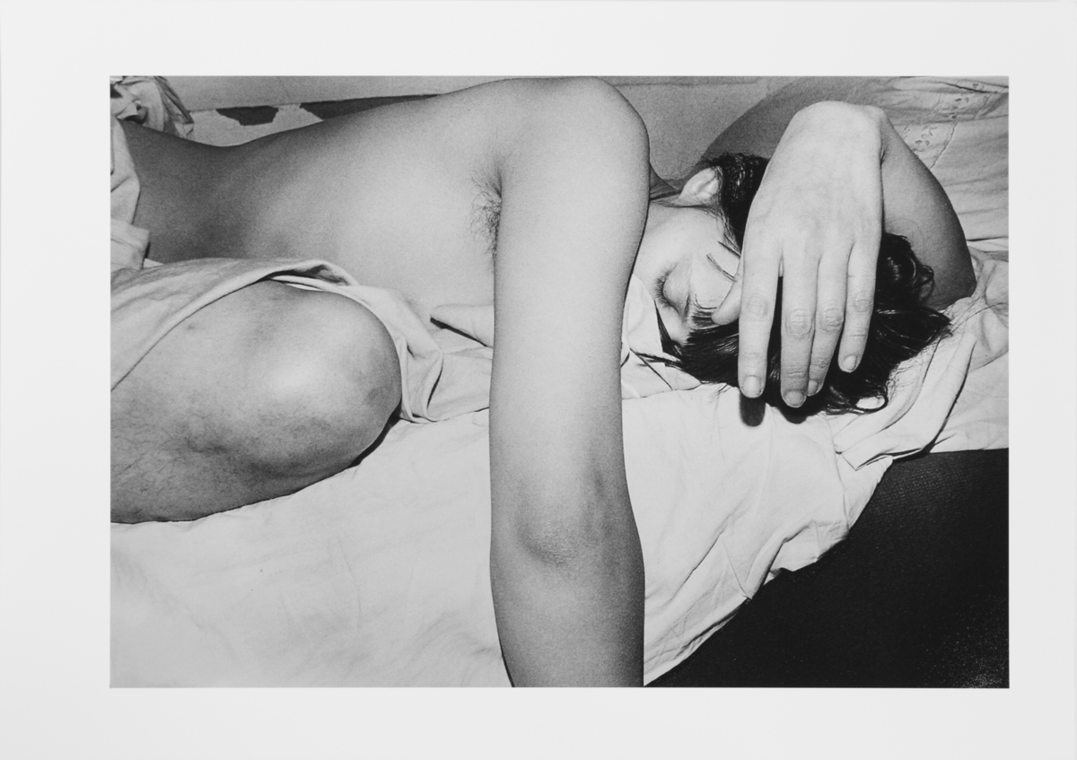 Konstnären liggandes på säng, med knäet uppdraget mot bröstet, högerarmen hängande längs sängkanten och vänster arm över huvudet. Halva kroppen synlig.