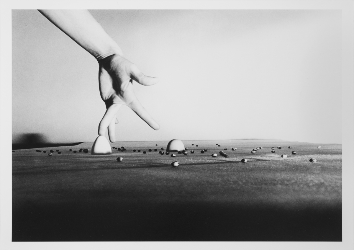 På ett bord är ett antal bönor utplacerade. Från vänster i bilden kommer delar av en arm och en hand där fingrarna promenerar över bordet.