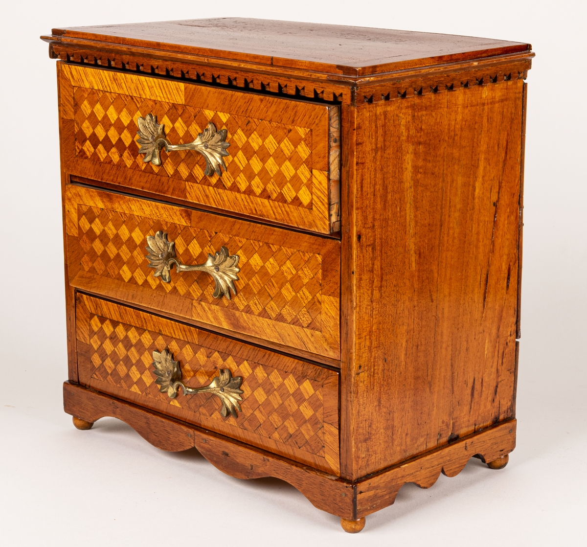 Sybyrå, barock typ, med tandning under skivans kanter. Miniatyrbyrå, med tre draglådor med fanér, delvis i rutmönster. Gjutna bronshandtag. Kulfötter. Obs. en liten smal låda upptill på byråns baksida.