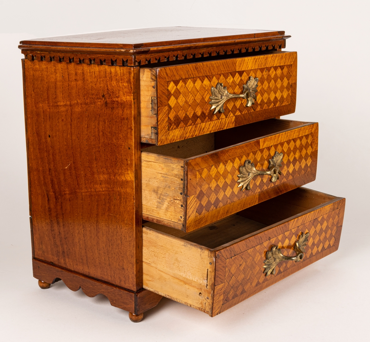 Sybyrå, barock typ, med tandning under skivans kanter. Miniatyrbyrå, med tre draglådor med fanér, delvis i rutmönster. Gjutna bronshandtag. Kulfötter. Obs. en liten smal låda upptill på byråns baksida.