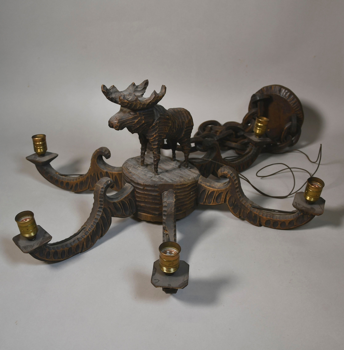 Lysekrone av tre, med seks armer. På midten er det en skåret figur av stående elg. På sidene av armene er det border med dekorative ellipser og sirkler, og en bølgeformet svai på toppen. Lysstellet henger i kjettinger av tre. Lampeskjermer av hvit tekstil er kjegleformet.