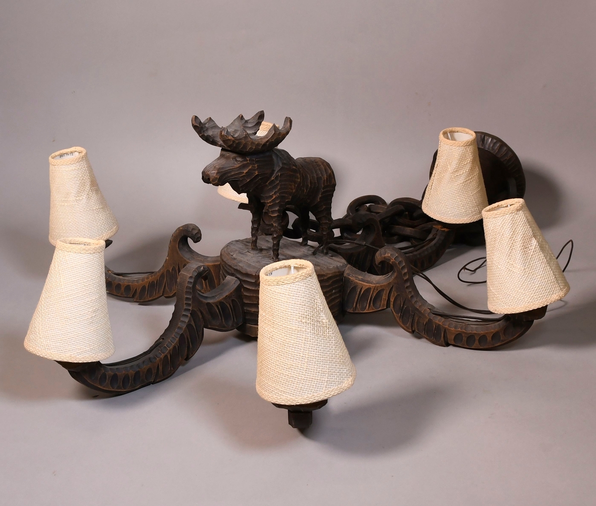 Lysekrone av tre, med seks armer. På midten er det en skåret figur av stående elg. På sidene av armene er det border med dekorative ellipser og sirkler, og en bølgeformet svai på toppen. Lysstellet henger i kjettinger av tre. Lampeskjermer av hvit tekstil er kjegleformet.