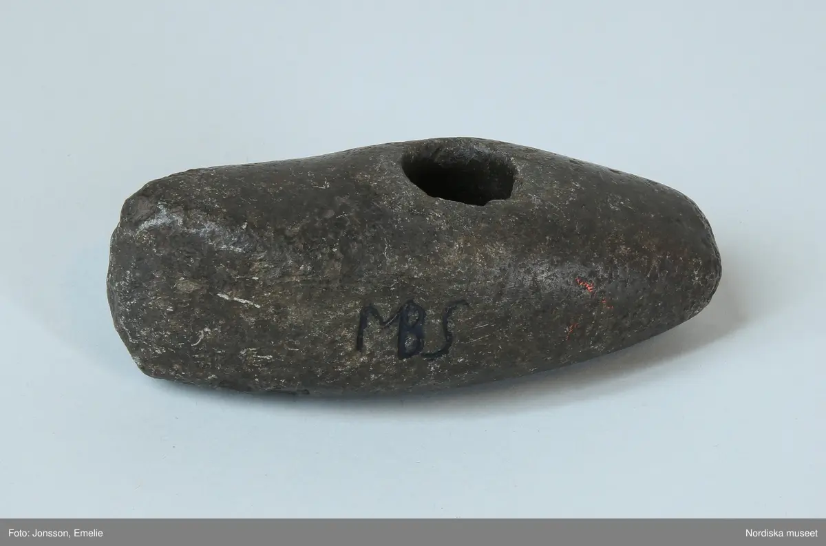 Huvudliggare:
"Stenyxa  fr. Södra Vi sn. Severeds hd. Småland.
'S.k. 'åskvigge', oval med skafthål, nästan trind med trubbig bane. Slipad. Kringskafthålet å framsidan en otydlig fals. 11 cm l., 4 cm. hög i eggen. Omkr. 4,5 cm br. öfver skafthålet.' Märkt: 'MBS'.
Eggen skadad. 
Har gått i arf i riksdagsman Nils Magnussons slägt i Södra Vi sn. och har nyttjats vid brännande af fällor på det sättet, att man dragit med den på marken en rispa som skulle ge tillkänna huru vidt elden skulle gå. Bokstäverna MBS betyder Magnus Bengtsson, namnet på riksdagsman Nils Magnussons fader, som helt säkert mäjslat in dessa bokstäver. Allt detta enl. uppgift af nu mera komministern i Eksjö Karl Viktor Villner, som skänkt yxan till undertecknad.' "

Bilaga:
Inget utöver huvudliggare

Ur "Svensk forntro och folksed i bild och ord", 1920, N.E.Hammarstedt, Stockholm. s. 17:
"ÅSK-VIGGE från Södra Vi kyrkby,  Södra Vi socken, Sevedes härad, Småland. N.M. 6,451. Borrad stenyxa, med på ena sidan inristat ägarmärke: MBS. Eggen avstött. L. 0,113 m. G. av aman. G. Norlander i Uppsala 1875.
Har gått i arv i riksdagsman Nils Magnussons släkt i Södra Vi socken och har nyttjats vid brännande av fällor på det sättet, att man dragit med den en rispa på marken, som skulle ge till känna, huru vitt elden skulle gå. Bokstäverna MBS betyda Magnus Bengtsson, riksdagsman Nils Magnussons fader, som helt säkert mäjslat in dessa bokstäver. Allt detta enligt uppgift av numera komministern i Eksjö C. V. Villner, som skänkt yxan till givaren. Nord. Mus. inv. kat."

Ur "Svensk forntro och folksed i bild och ord", 1920, N.E.Hammarstedt, Stockholm. s. 14:
"ÅSK-VIGGAR
ÅSK-VIGGAR, tor-viggar eller tor-blegar kallas av allmogen i åtminstone de flesta av våra landskap i jorden funna stenåldersredskap, företrädesvis stenyxor, såväl borrade som även oborrade. men även andra om dylika mer eller mindre erinrrande naturformade stenar erhålla ofta denna benämning. Det troddes, att en sådan åskvigg nedföll vid varje åskslag, men nedträngde så djupt (sju alnar eller sju famnar) i jorden, att den först efter flera (vanligen sju) år kom i dagen. Dylika åskviggar tillskriver man ännu i åtskilliga orter stor kraft i flera hänseenden. Ofta skrapade man även pulver av dem för att använda det som läkemedel (jfr älvkvarnar och älvstenar)."
