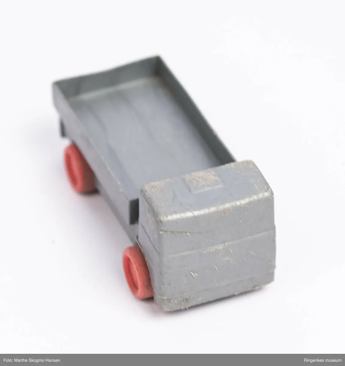 Liten lekebil av plastikk. Bilen er formet som en lastebil med åpent lasteplan. Fronten er formet slik at det vagt ligner karosseri og vindu. Hjulene er av rød plast, og sitter på rustne metall-akslinger.