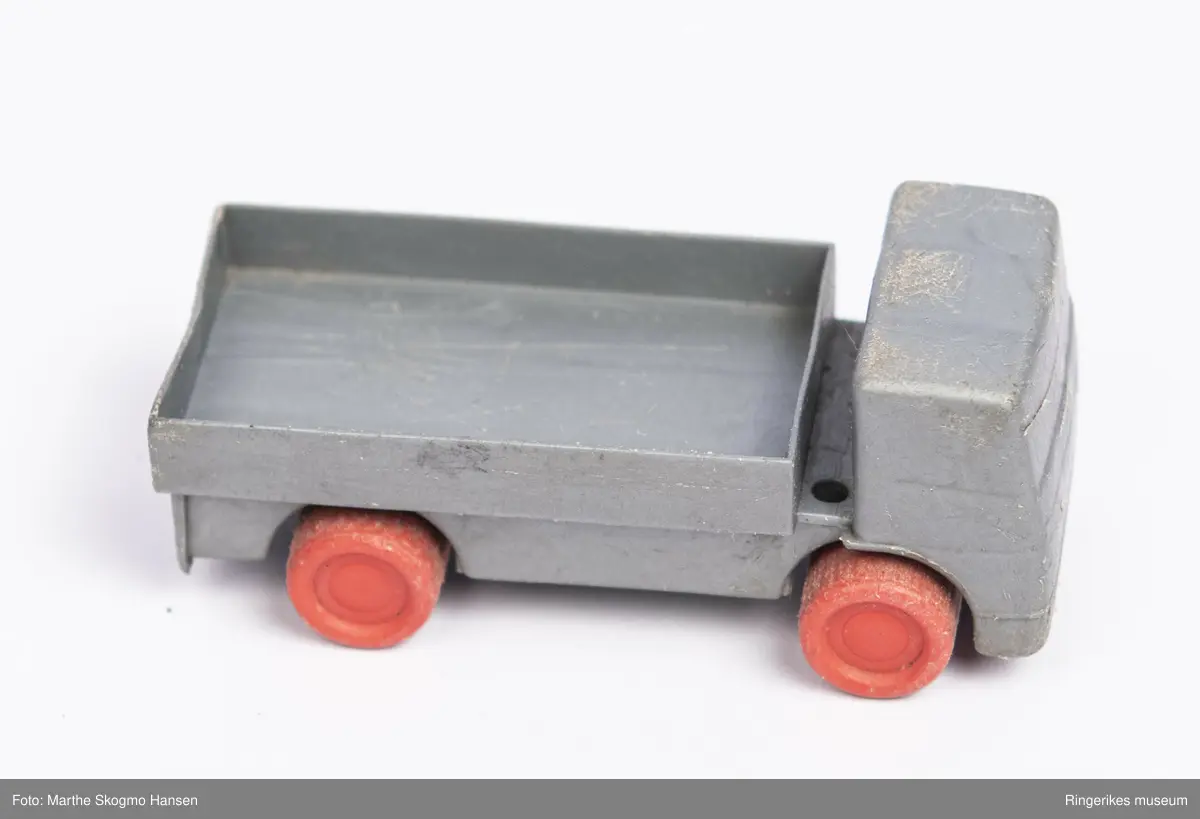 Liten lekebil av plastikk. Bilen er formet som en lastebil med åpent lasteplan. Fronten er formet slik at det vagt ligner karosseri og vindu. Hjulene er av rød plast, og sitter på rustne metall-akslinger.