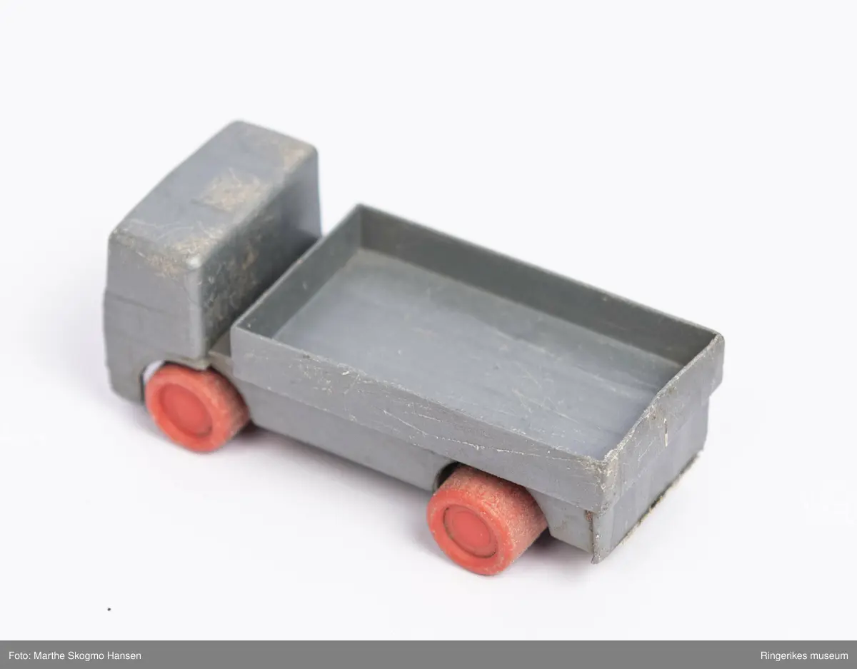 Liten lekebil av plastikk. Bilen er formet som en lastebil med åpent lasteplan. Fronten er formet slik at det vagt ligner karosseri og vindu. Hjulene er av rød plast, og sitter på rustne metall-akslinger.